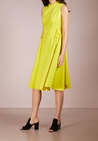 Žluté bezrukávové šaty s kulatým výstřihem a detailem na stažení, doplněné černými otevřenými blokovými podpatky na hladkém povrchu.