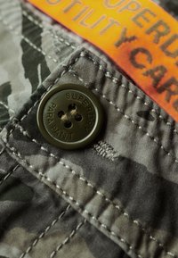 Utilitybroek van camouflage-stof met een donkergroene knoop waarop "SUPERDRY PARA PANT" is gegraveerd. Een feloranje label is zichtbaar.