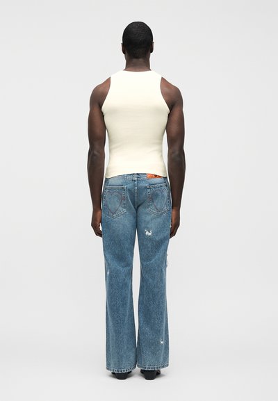 Μπεζ αμάνικη μπλούζα και ανοιχτόχρωμα τζιν παντελόνια με φαρδύ πόδι και φθαρμένες λεπτομέρειες. Τα τζιν έχουν τσέπες στην πλάτη σε σχήμα καρδιάς με κόκκινη ραφή.