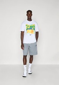 Camiseta blanca con un estampado gráfico colorido, pantalones cortos grises, zapatillas blancas y calcetines blancos, con un logo negro de Nike en la camiseta y en los pantalones cortos.