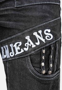 Close-up of black denim jeans pocket with white embroidered text "JEANS" and silver rivet details on vertical straps.