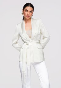 Femme aux cheveux bruns portant un blazer blanc ceinturé avec des détails à franges, un haut beige, un pantalon blanc et des boucles d'oreilles dorées remarquables sur un fond uni.