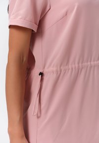Rosa Bluse aus einem glatten, leichten Stoff. Sie hat kurze, umgeschlagene Ärmel, einen elastischen Bund und eine Kordel mit einem schwarzen Stopper.