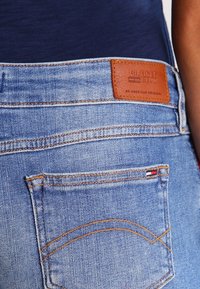 Vaatteiden takapuoli vaaleansinisestä denimistä, ruskea nahkainen Hilfiger Denim -merkki ja pieni punaisen, valkoisen ja sinisen värinen lippu taskussa.