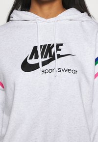 Sudadera ligera gris claro con el logo negro de Nike y el texto "sportswear". Presenta rayas coloridas en rosa, verde y azul en las mangas.