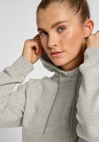 Grå hoodie med snörning, mjukt material och ribbade manchetter. Modellen justerar huvan och visar den avslappnade passformen och texturen.