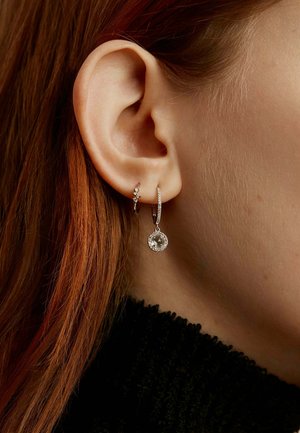Pendientes de aro de plata con un colgante de gema clara; un pendiente presenta un pequeño acento de piedra. Llevados en la oreja izquierda contra el cabello rojo.