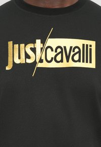 Juoda medvilninė marškinė su ryškiu auksiniu tekstu "just/cavalli", pasižyminčia kontrastingomis tekstūromis ir apvaliu iškirpimu.
