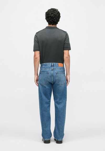 Sötétszürke, rövid ujjú pólóing, amely világoskék, laza szabású farmernadrággal van párosítva. A derékpánton bőrből készült folt található.