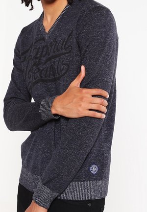 Mand iført en mørkegrå V-hals sweater med teksten "Leopard Jeans" tværs over brystet og et lille rundt logo nær kanten.