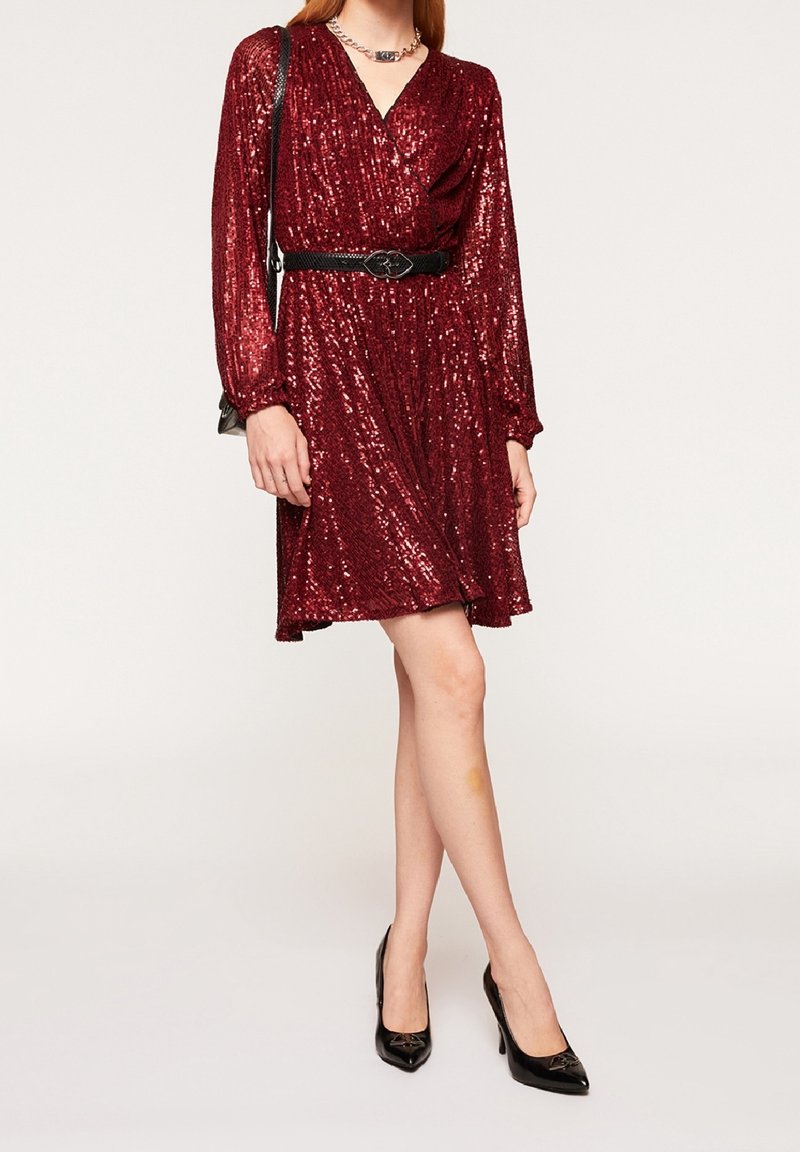 Robe en sequins bordeaux avec des manches longues, design enroulé, et une ceinture noire. Associée à des escarpins noirs à bout pointu, créant un effet scintillant.