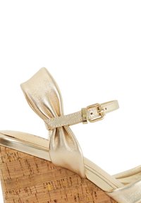 Gouden wig sandaal met een metallic afwerking, kurken platform en een strikaccent op de enkelband, met een gesp-sluiting.