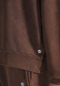 Felpa marrone con texture a coste e polsini, caratterizzata da una piccola patch con logo vicino all'orlo. Design semplice, a taglio dritto, con una vestibilità rilassata.