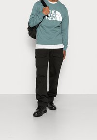 Man som bär en teal The North Face-tröja, svarta cargobyxor, svarta skor och bär en svart ryggsäck över ena axeln.