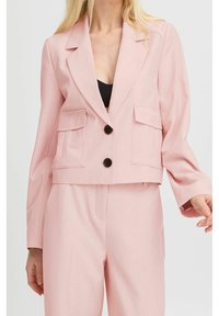 Světle růžové zkrácené sako z texturované tkaniny, vybavené dvěma předními kapsami a dvěma černými knoflíky na zapínání.