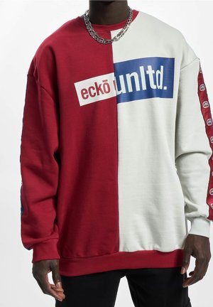 Personne portant un sweat-shirt bicolore rouge et blanc avec le texte « eckō unltd. », une bande logo sur les manches et un collier chaîne argenté.