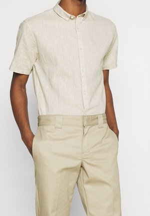 Homme portant une chemise beige clair à manches courtes boutonnée rentrée dans un pantalon beige taille haute, les mains dans les poches.