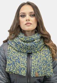Sciarpa di maglia blu e gialla con un pattern testurizzato, appoggiata attorno al collo, abbinata a un giubbotto invernale grigio.