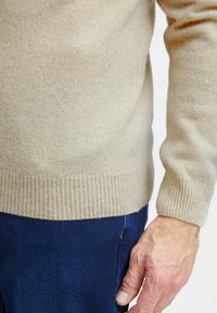 Beige kasmír pulóver texturált kötésű, bordázott mandzsettákkal és szegéllyel; sötétkék nadrággal párosítva, melyen egy oldalzseb látható.