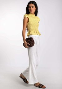Top amarillo con fruncidos y dobladillo con volantes, combinado con pantalones anchos blancos y sandalias negras de tiras, sosteniendo un bolso de mano marrón texturizado.
