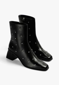 Botines negros hasta el tobillo con puntera cuadrada, hechos de cuero liso, con tachuelas metálicas dispersas por toda la superficie y tacón cuadrado.