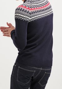 Person som bär en marinblå tröja med vitt och rött mönstrat ok och mörka jeans, visad bakifrån med händerna ihopknäppta.