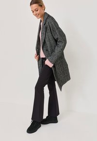 Grau texturierter Mantel, rosa Pullover, schwarze weit geschnittene Hose und schwarze Sneakers. Der Mantel hat eine lockere Passform mit einem Reverskragen und Knöpfen.