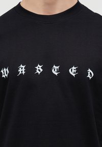 Άνθρωπος που φοράει μαύρο πουκάμισο με λευκά, στυλιζαρισμένα γοτθικά γράμματα που σχηματίζουν τη λέξη "WASTED" στο στήθος.