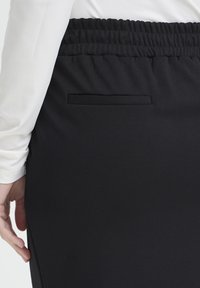 Osoba nosící černou sukni s elastickým pasem, zadní kapsou s rozparkem a bílým dlouhým rukávem, stojící s levou rukou volně podél těla.