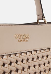 Beige vævet håndtaske med glat læderoverdel, guldtonet logo "GUESS EST 1981" og dekorativ syning langs kanterne.