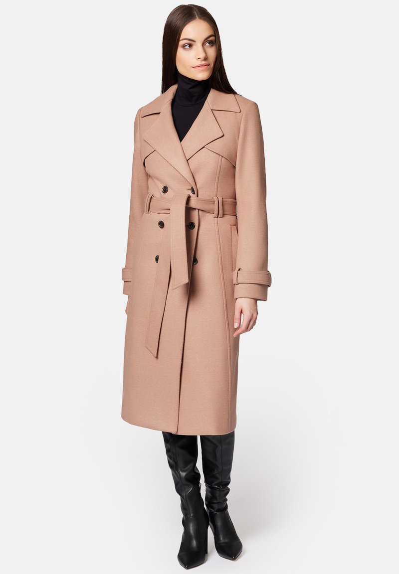 Beżowy wełniany płaszcz z podwójnym zapięciem, paskiem w talii, klapami i bocznymi kieszeniami. Połączony z czarnymi botkami do kolan.