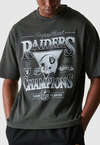 Personne portant un t-shirt noir et gris des Raiders Champion du Super Bowl 1983 avec un casque de football et le logo de la NFL.