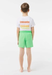 Witte T-shirt met meerkleurige "Lacoste" tekst op de achterkant; groene zwembroek met elastische tailleband, gladde textuur en boven de knie lengte.