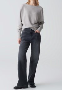 Kobieta ubrana w jasnoszary sweter z długim rękawem i ciemne, szerokie dżinsy z czarnymi butami, stojąca z jedną ręką w kieszeni na jednolitym tle.