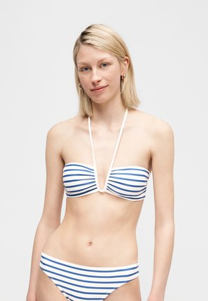 Blondínka v modro-bielom pruhovanom bikinách s remienkom za krkom, stojaca na jednoduchom svetlosivom pozadí.