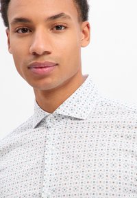 Camicia bianca con un delicato motivo floreale in verde e beige, con colletto classico e chiusura con bottoni. Vista ravvicinata del tessuto e del design.