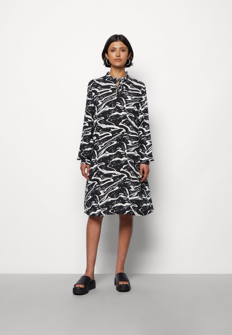 Vestido em mármore preto e branco com colarinho redondo, mangas longas e detalhe de laço no pescoço. Modelo está de sandálias de plataforma pretas.
