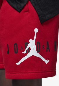 Pantalones cortos rojos de tela suave con un gran logo blanco de "Jumpman" y texto negro "JORDAN". La cintura es elástica con un acabado suave.
