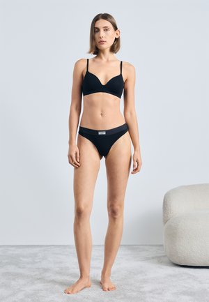 Soutien-gorge noir et culotte assortie en tissu lisse, présentant un décolleté plongeant et une étiquette discrète à la taille. Le modèle se tient pieds nus sur un tapis.