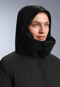 Crna jakna s kapuljačom i visokim ovratnikom, izrađena od glatkog, vodootpornog materijala. Uključuje nabrane šavove oko područja lica i vrata.