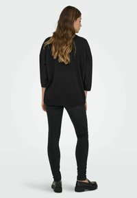 Mujer con cabello largo y castaño, vestida con un suéter negro suelto, jeans negros ajustados y mocasines negros, de pie de espaldas sobre un fondo blanco.