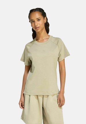 Frau trägt beige kurzärmliges T-Shirt und passende lockere Shorts, steht mit entspannter Haltung vor einfarbigem Hintergrund.