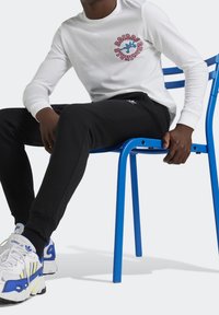 Bílá košile s dlouhým rukávem a barevným potiskem, černé tepláky a bílé tenisky s modrými a žlutými akcenty, sedící na modré židli.