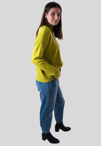 Vrouw in een groene trui en blauwe jeans die zijwaarts staat, met zwarte hoge hakken op een effen lichtgrijze achtergrond.