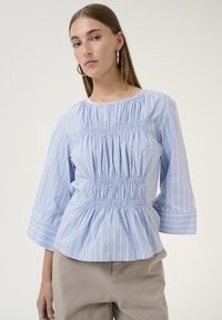 Blusa blu a righe con maniche a sbuffo e dettagli arricciati in vita. Tessuto di cotone liscio, caratterizzato da righe bianche verticali.