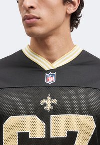 Άνδρας φορώντας μαύρη και χρυσή φανέλα New Orleans Saints NFL με τον αριθμό 27 και το λογότυπο fleur-de-lis μπροστά.
