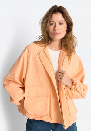 Žena s vlnitými svetlohnedými vlasmi, oblečená v voľnej broskyňovej zipovej bunda cez biele tričko a modré džínsy, pózuje pred jednoduchým bielym pozadím.