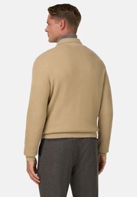 Beige strukturstickad tröja med ribbad yta. Har en krage och är ihopparad med mörkgrå byxor. Bakifrån visar designen.