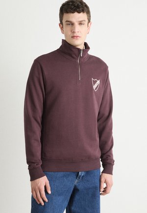 Maroon quarter-zip sweatshirt with ribbed cuffs and hem. Features a small, white shield logo on the chest. Soft, textured fabric.