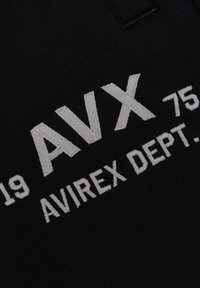 Zwarte stof met witte geborduurde tekst "AVX 75 AVIREX DEPT." Het ontwerp heeft opvallende, vetgedrukte letters en een gestructureerde uitstraling.